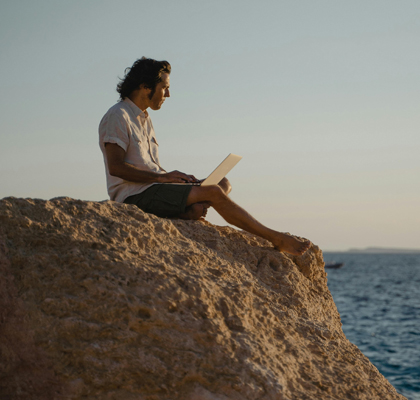 Ein Mann sitzt mit Laptop auf einem Felsen am Meer