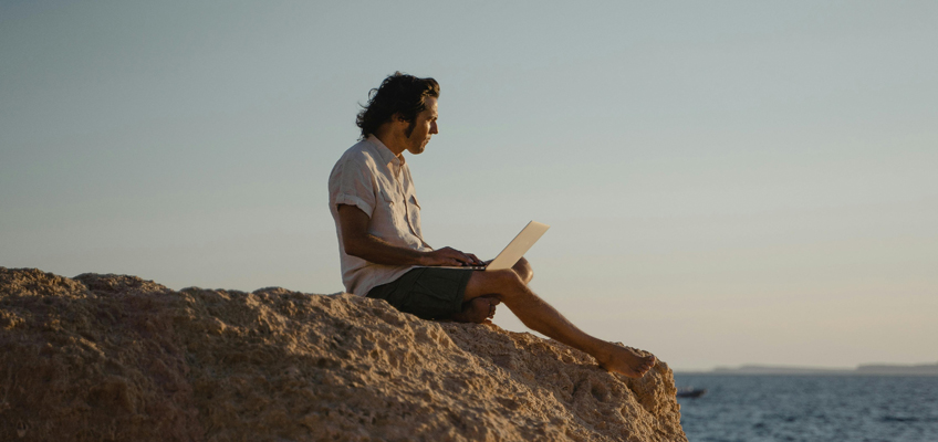 Ein Mann sitzt mit Laptop auf einem Felsen am Meer